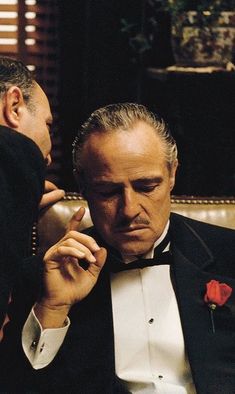 a man in a tuxedo sitting next to another man with a red rose on his lapel