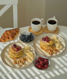 two plates with pancakes, bananas and berries on them sitting next to cups of coffee