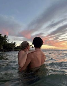 a man and woman are sitting in the ocean watching the sun set over the water