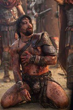 a man with no shirt sitting on the ground in front of other men wearing armor