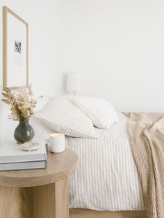 a bed with white sheets and pillows next to a table with a vase on it