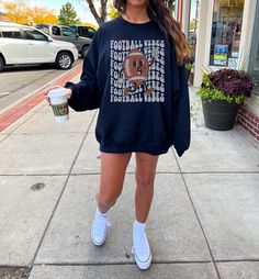 a woman is walking down the sidewalk holding a coffee cup and wearing a football sweatshirt