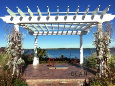 a white pergolated area next to the ocean with flowers and plants around it