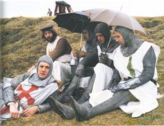 a group of men dressed in medieval clothing sitting on the grass with an umbrella over their heads