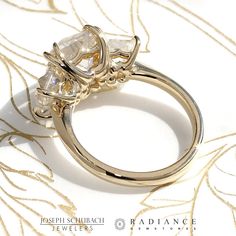 three stone ring sitting on top of a white and gold brocaded cloth with the words radiance written below it