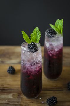 two glasses filled with blackberries and ice on top of a wooden table next to berries