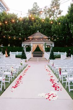 an outdoor ceremony setup with white chairs and red rose petals on the aisle, surrounded by string lights