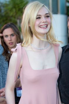 a woman with blonde hair wearing a pink dress and smiling at the camera while standing in front of other people