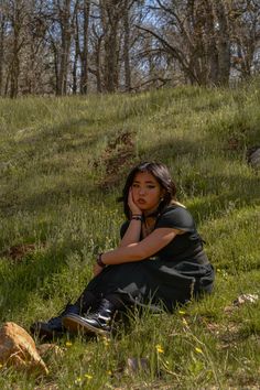 a woman sitting in the grass with her hand on her chin and looking off into the distance