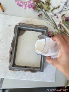 someone is holding a glass in front of a picture frame with flowers on the table