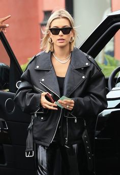 a woman in black jacket and leather pants standing next to a car with the door open