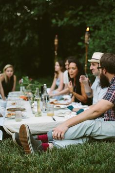 a group of people sitting at a picnic table eating food and drinking wine in the park