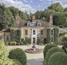 a large brick house surrounded by lush green trees and bushes with a fountain in the middle