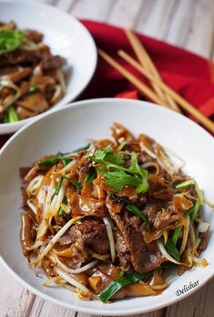 two white bowls filled with beef and noodles on top of a wooden table next to chopsticks