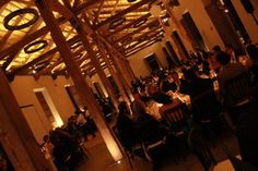 people are sitting at tables in a large room with wooden beams on the ceiling and lights hanging from the ceiling