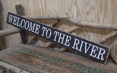 a welcome to the river sign sitting on top of a wooden bench next to a wall