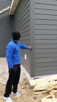 a man in blue shirt and black pants standing next to a gray house with siding on it