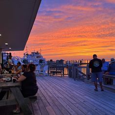 people are sitting at picnic tables on the dock as the sun sets over the water