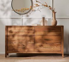 a wooden dresser with a mirror and vase on it in front of a white wall