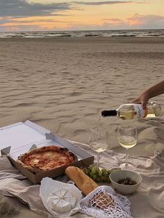 a person pouring wine into a pizza box on the beach with other food and drinks