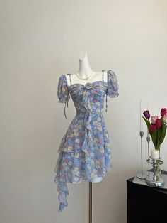 Circuitous and crafted with care, this blue floral dress weaves its way through timeless fashion with a modern twist. The layers of lightweight fabric create a gentle cascade, giving this dress a buoyant, ethereal quality that’s perfect for spring and summer events. Decorated with a vibrant array of floral patterns in soft hues, the dress features dainty shoulder straps and a rosette detail for a touch of romance. Its asymmetrical hemline and flowing silhouette ensure it moves gracefully with th Asymmetrical Dress Pattern, Cascade Dress, Floral Cascade, Victorian Dresses, Wardrobe Goals, Blue Floral Dress, Clothing Pieces, Old Hollywood Glamour, Floral Blue Dress