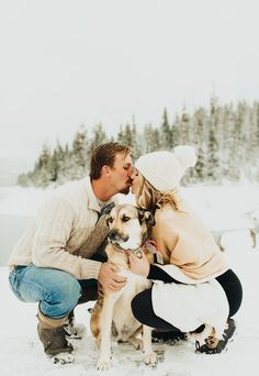 a man and woman kissing their dog in the snow with trees in the back ground