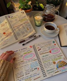 a person is writing in a notebook next to a cup of coffee and an open book