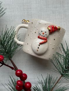 a white mug with a snowman on it sitting next to some christmas tree branches