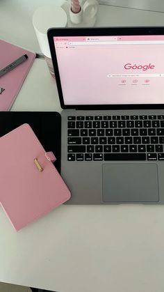 an open laptop computer sitting on top of a white desk next to a pink notebook