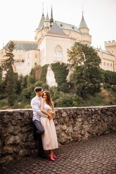 a man and woman standing next to each other in front of a castle on top of a hill