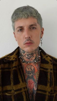 a man with tattoos and piercings on his chest wearing a jacket over a white wall