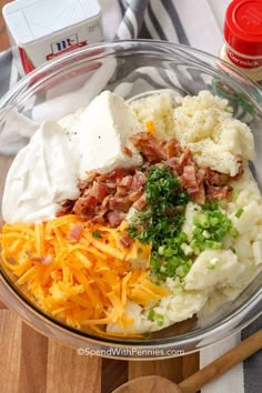 a glass bowl filled with mashed potatoes, cheese and bacon on top of a wooden cutting board