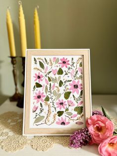 a card with pink flowers and green leaves in front of two candles on a lace doily