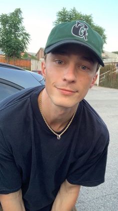 a young man wearing a green hat sitting in front of a black car with his hands on his hips