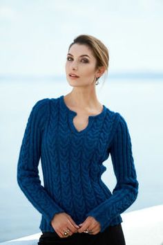 a woman wearing a blue sweater standing next to the ocean with her hands in her pockets