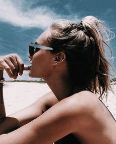 a woman sitting on top of a sandy beach next to the ocean with her finger in her mouth