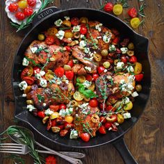 a skillet filled with chicken, tomatoes and other vegetables on top of a wooden table