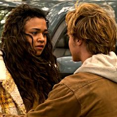 two people standing next to each other in front of a car and one person with long hair