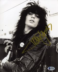 a black and white photo of a man with long hair wearing a leather jacket, standing in front of a fence