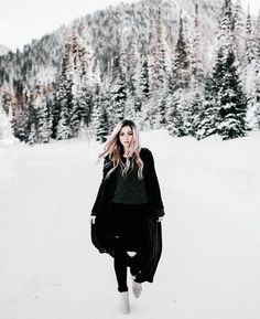 a woman is walking in the snow with her hair blowing in the wind and trees behind her