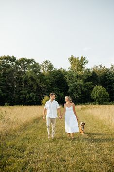 a man and woman holding hands while walking their dog through the grass with trees in the background