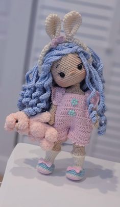 a crocheted doll with blue hair holding a pink teddy bear in her arms