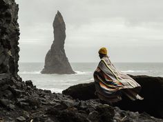 a person wrapped in a blanket sitting on rocks near the ocean with an island in the background