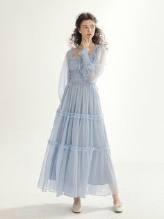 This product is an Isabel Smock Long Dress that exudes romantic elegance with its tiered tulle skirt and delicate smocking detail. The long sleeves are complemented by the smocked bodice, offering a snug fit with a vintage flair. This dress combines the airy lightness of the fabric with the intricate detail of the smocking, creating a harmonious blend of sophistication and whimsy. - The dress is adorned with tiered tulle layers, each accented with a ruffle trim, adding dimension and a fairy-tale quality.- Its smocked bodice provides both comfort and a tailored appearance, enhancing the dress's feminine appeal.- The long, sheer sleeves offer coverage while maintaining the dress's ethereal aesthetic.- Crafted for those special occasions, the Isabel Smock Long Dress is a beautiful choice Light Blue Cottagecore Dress, Ethereal Dress Casual, Elegant Tiered Smocked Dress With Ruched Detail, Elegant Ruched Tiered Smocked Dress, Elegant Long Sleeve Smocked Dress With Smocked Cuffs, Elegant Long Sleeve Smocked Dress, Elegant Smocked Dress With Ruffles And Tiered Skirt, Feminine Smocked Dress With Long Sleeves, Elegant Long Sleeve Flowy Smocked Dress