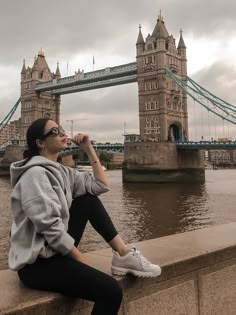 a woman sitting on the edge of a wall next to a river with a bridge in the background