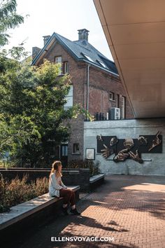 a woman sitting on top of a bench next to a brick wall and tree covered building