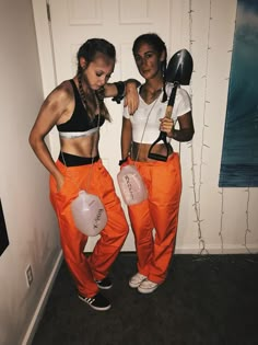 two young women dressed in orange pants and white shirts, one holding a pair of scissors