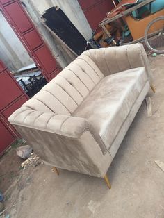 a tan couch sitting on top of a dirty floor next to a red building with lots of junk