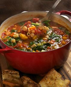 a red pot filled with vegetable soup next to some bread on a wooden cutting board