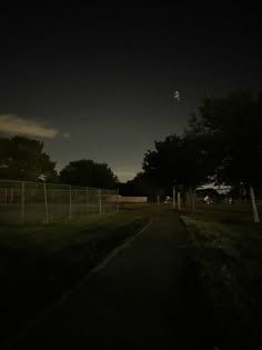 an empty road at night with the lights on and trees in the background, along with a fence